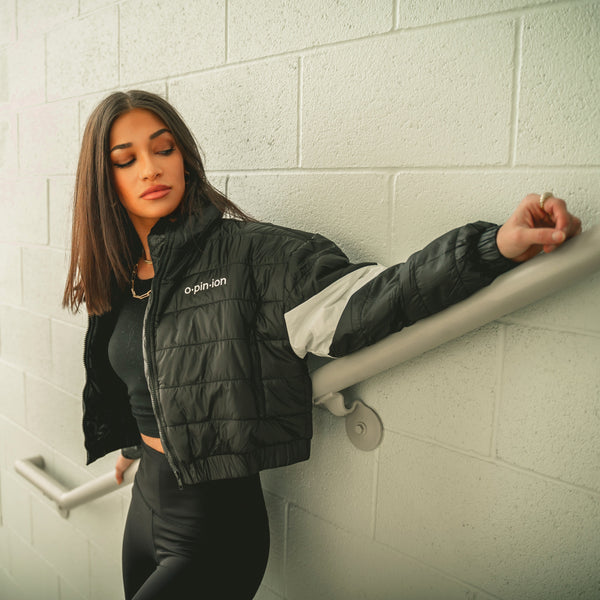 Woman in a black puffer jacket standing on a staircase with a white tiled wall.
