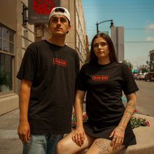 Load image into Gallery viewer, Two people wearing black t-shirts with a red logo on a city street.