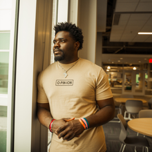Load image into Gallery viewer, Man wearing a beige t-shirt with text, standing indoors near a glass door.
