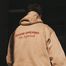 Load image into Gallery viewer, Person wearing a beige hoodie with 'CHASE DREAMS Not Approval' text on the back against a dark background