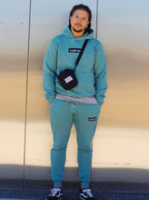 Load image into Gallery viewer, Person wearing a matching teal hoodie and jogger set with a black crossbody bag against a metallic wall.
