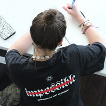 Load image into Gallery viewer, Person wearing a black t-shirt with a logo, sitting at a desk and writing on a piece of paper.